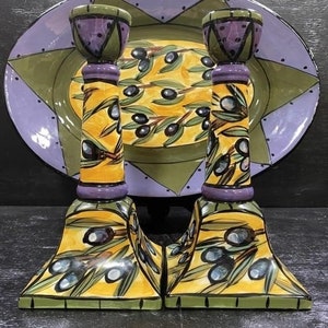 May include: A set of two ceramic candle holders with an olive branch design. The holders are yellow, green, and purple with a detailed olive branch pattern. The holders are sitting on a black surface.