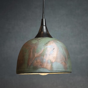 May include: A pendant light fixture with a dome-shaped shade. The shade has a mottled green and copper finish, with a black metal top and a single light bulb visible at the bottom. The fixture hangs from a black cord.