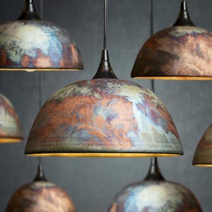 May include: Several pendant lights with a unique, dome-shaped design. The lampshades feature a mottled, earthy color palette of copper, blue, and cream. Each light hangs from a black cord and fixture, creating a warm, ambient glow.