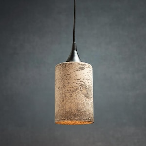 May include: A cylindrical pendant light fixture featuring a textured, mottled beige and brown exterior finish. The lamp is suspended by a thin black cord attached to a smooth, conical black metal cap at the top, with a warm light emitting from below.