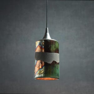 May include: A cylindrical pendant light fixture featuring a glossy, multi-colored glazed ceramic shade with iridescent green, copper, and dark tones. A matte grey horizontal band wraps around the center, suspended by a thin black cord.