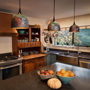 May include: Three dome-shaped pendant lights with a dark, multi-coloured finish featuring swirling patterns of green, blue, and copper tones. Each light hangs from a thin black cord, suspended above a kitchen island with a dark countertop.