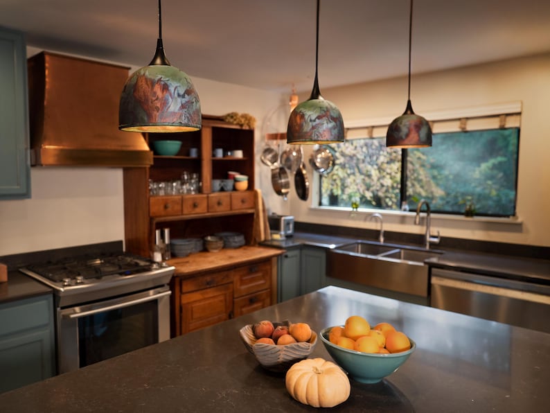 May include: Three dome-shaped pendant lights with a dark, multi-coloured finish featuring swirling patterns of green, blue, and copper tones. Each light hangs from a thin black cord, suspended above a kitchen island with a dark countertop.