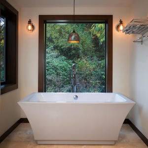 May include: A white rectangular freestanding bathtub with a chrome overflow drain, positioned in front of a large window. The bathroom features two wall-mounted light fixtures with exposed bulbs, a hanging pendant light, and a chrome towel rack.