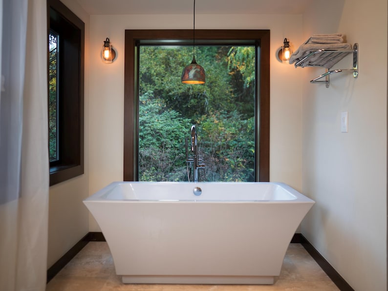 May include: A white rectangular freestanding bathtub with a chrome overflow drain, positioned in front of a large window. The bathroom features two wall-mounted light fixtures with exposed bulbs, a hanging pendant light, and a chrome towel rack.