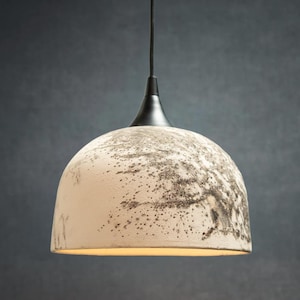 May include: A dome-shaped pendant light fixture with a textured off-white surface featuring dark gray speckled patterns. The lamp has a smooth black metal cap at the top where it connects to a thin black cord, casting a warm glow from underneath.