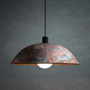 May include: A pendant light fixture with a copper-colored, dome-shaped shade. The shade has a mottled, textured appearance with shades of red, blue, and brown. A white light bulb is visible at the bottom. The fixture hangs from a black cord.