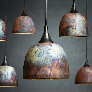 May include: A collection of dome-shaped pendant lights featuring a mottled, multi-coloured finish with swirls of copper, blue, and earthy brown tones. Each lamp is suspended by a black cord and topped with a dark metal fixture, creating a cluster.