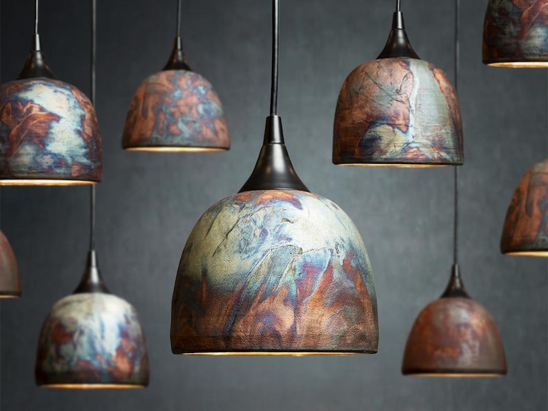 May include: A collection of dome-shaped pendant lights featuring a mottled, multi-coloured finish with swirls of copper, blue, and earthy brown tones. Each lamp is suspended by a black cord and topped with a dark metal fixture, creating a cluster.