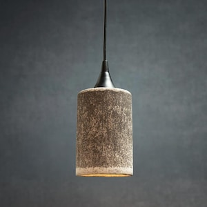 May include: A cylindrical pendant light fixture featuring a textured, mottled brown and gray ceramic shade. The shade is suspended by a thin black cord attached to a black metal cap at the top, with a warm light emitting from the bottom opening.