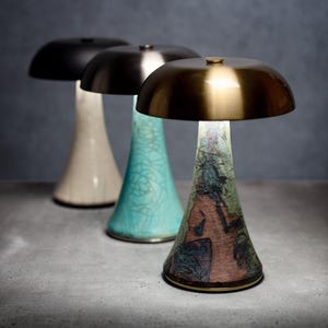 May include: Three mushroom-shaped table lamps with metallic dome shades. The front lamp features a textured, multi-colored base with green and brown tones. The middle lamp has a light blue crackle-patterned base, and the back lamp has a cream base.