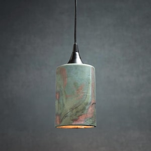 May include: A cylindrical pendant light fixture featuring a textured ceramic shade with a mottled green and reddish-brown glaze. The shade is suspended by a black cord attached to a black metal cap at the top, with a warm light emitting from the base.