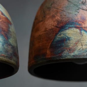 May include: Two dome-shaped ceramic pendant lights featuring a textured, multi-coloured glaze. The surface displays a blend of deep copper, burnt orange, and metallic blue tones with a rough, matte finish and visible cracks across the curved exterior.