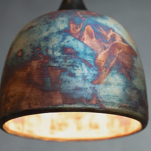 May include: A dome-shaped pendant light fitting featuring a textured ceramic surface with a marbled finish in shades of deep blue, copper, and earthy brown. The bottom opening reveals a bright, warm light source emanating from within the shade.