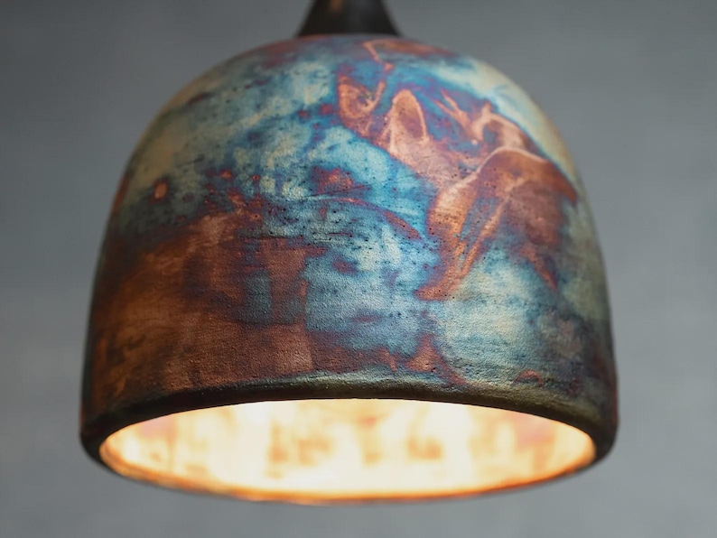 May include: A dome-shaped pendant light fitting featuring a textured ceramic surface with a marbled finish in shades of deep blue, copper, and earthy brown. The bottom opening reveals a bright, warm light source emanating from within the shade.