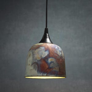 May include: A dome-shaped pendant light fixture featuring a ceramic shade with a mottled, multi-colored glaze in shades of copper, blue, and cream. The shade is attached to a black metal cap and a black cord, with a warm light emitting from below.