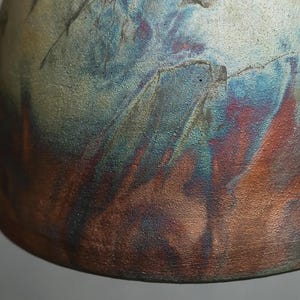 May include: A ceramic vessel with a textured surface featuring a metallic glaze in shades of deep blue, copper, and bronze. The finish displays an iridescent quality with dark, smoky streaks and a rough, granular appearance across the curved body.