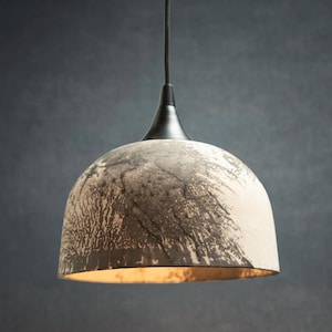 May include: A dome-shaped pendant light fixture featuring a textured, mottled gray and off-white exterior finish. The lamp is suspended by a thin black cord attached to a smooth, conical black metal cap at the top, with a warm light glowing from within.