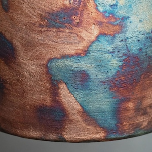 May include: A cylindrical object with a textured surface featuring a metallic copper finish. The surface displays irregular, mottled patches of oxidised blue and dark purple tones, creating a variegated appearance across the curved exterior.