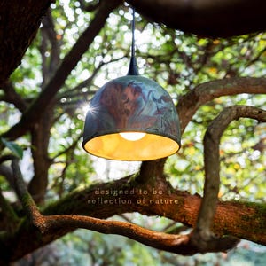 May include: A dome-shaped pendant light with a mottled blue and copper patina finish on the exterior and a warm gold interior. The light features a black cord and socket, with the text "designed to be a reflection of nature" printed below.
