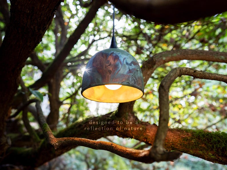 May include: A dome-shaped pendant light with a mottled blue and copper patina finish on the exterior and a warm gold interior. The light features a black cord and socket, with the text "designed to be a reflection of nature" printed below.