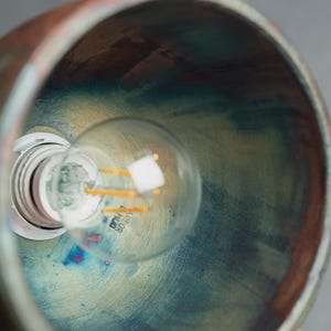May include: A bowl-shaped pendant light fitting with a metallic copper exterior and an interior featuring a blended finish of iridescent blue, green, and gold tones. A clear glass light bulb with visible filaments is installed in the centre socket.