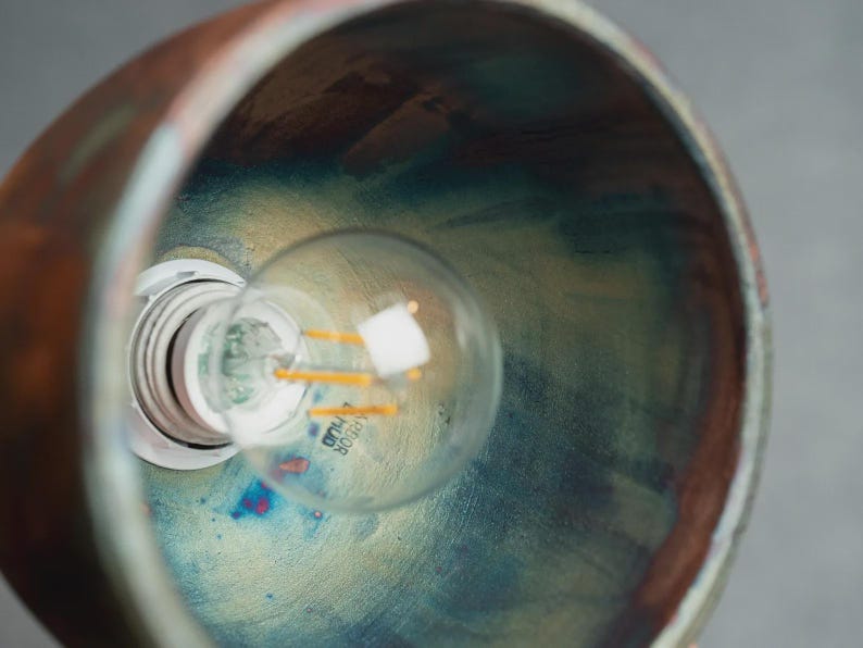 May include: A bowl-shaped pendant light fitting with a metallic copper exterior and an interior featuring a blended finish of iridescent blue, green, and gold tones. A clear glass light bulb with visible filaments is installed in the centre socket.