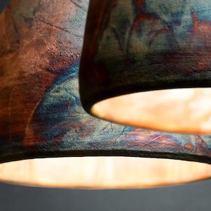 May include: Two dome-shaped pendant lights with a textured, rustic finish. The exterior surfaces feature a blend of deep copper, bronze, and iridescent blue tones. The interior of each shade is illuminated, casting a warm, soft glow downwards.