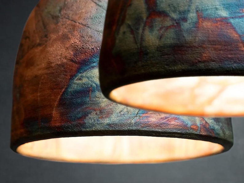 May include: Two dome-shaped pendant lights with a textured, rustic finish. The exterior surfaces feature a blend of deep copper, bronze, and iridescent blue tones. The interior of each shade is illuminated, casting a warm, soft glow downwards.
