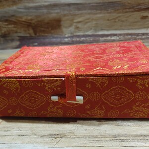 May include: A red box with a gold pattern and a white clasp. The box is closed and has a traditional Chinese design.