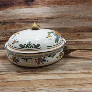 May include: White enamel cookware with a floral design in shades of blue, yellow, and green. The pot has a brass handle and a lid with a brass knob.