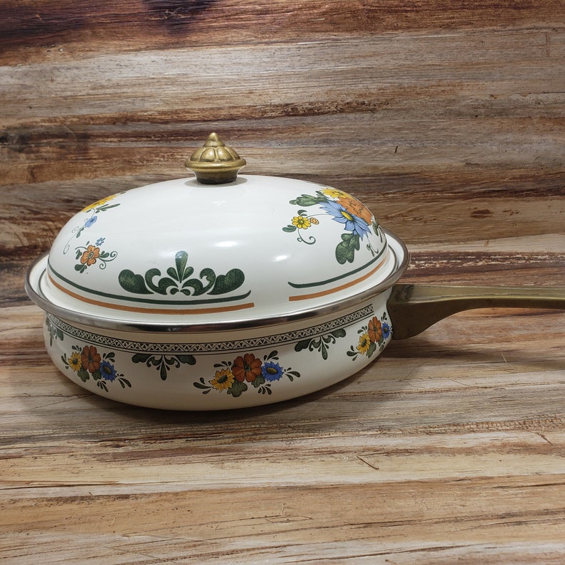 May include: White enamel cookware with a floral design in shades of blue, yellow, and green. The pot has a brass handle and a lid with a brass knob.