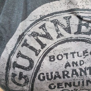 K&ouml;nnte beinhalten: Nahaufnahme eines schwarzen T-Shirts mit einer grauen und wei&szlig;en Grafik des Guinness-Logos. Das Logo lautet "Guinness Bottled and Guaranteed Genuine".