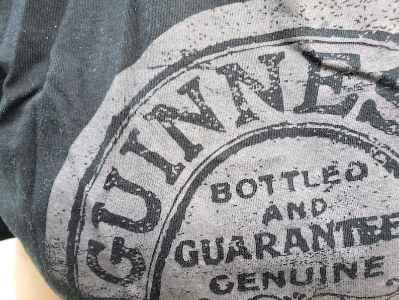 K&ouml;nnte beinhalten: Nahaufnahme eines schwarzen T-Shirts mit einer grauen und wei&szlig;en Grafik des Guinness-Logos. Das Logo lautet "Guinness Bottled and Guaranteed Genuine".