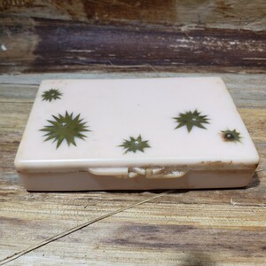 May include: A vintage pink plastic trinket box with a hinged lid. The box has a starburst design with gold accents and a small clear rhinestone in the center of each star.
