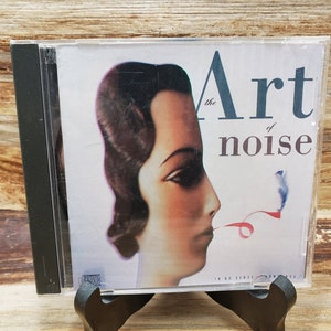 May include: CD case for the album "The Art of Noise." The cover features a woman's profile with a red ribbon and the album title in a stylized font. The CD case is in a clear plastic case.