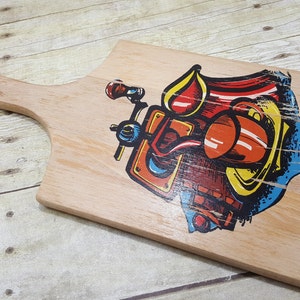 May include: A wooden cutting board with a colorful illustration of a vintage car. The board has a leather handle and is perfect for serving cheese, crackers, or other snacks.