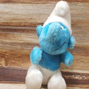 May include: A blue and white plush toy of a Smurf character. The Smurf is wearing a white hat and has a blue body with a white belly. The toy is sitting on a wooden surface.