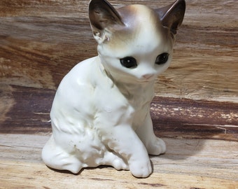 May include: A white ceramic figurine of a kitten with brown markings on its head and ears. The kitten is sitting with its head tilted down and its eyes closed.