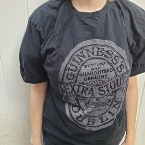 K&ouml;nnte beinhalten: Schwarzes T-Shirt mit einem grauen Aufdruck des Guinness-Logos. Das Logo lautet "Bottled and Guaranteed Genuine Extra Stout Dublin Ireland".