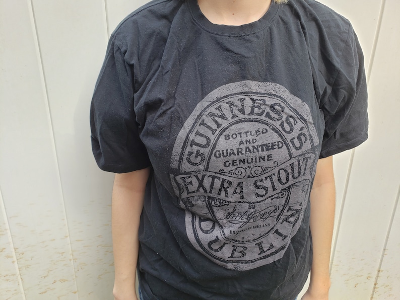 K&ouml;nnte beinhalten: Schwarzes T-Shirt mit einem grauen Aufdruck des Guinness-Logos. Das Logo lautet "Bottled and Guaranteed Genuine Extra Stout Dublin Ireland".