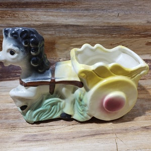 May include: A ceramic planter shaped like a horse pulling a wagon. The horse is black and white with brown accents. The wagon is yellow with a pink wheel. The planter is a vintage style.