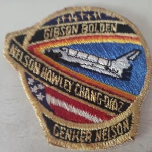 May include: A gold embroidered patch with a space shuttle flying over a blue and orange background. The text on the patch reads "Gibson Bolden, Nelson Hawley Chang-Diaz, Cenker Nelson".