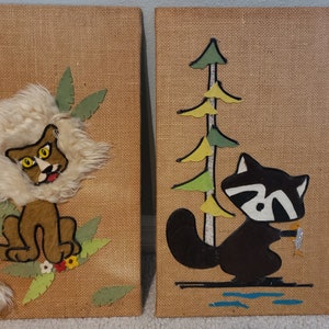 May include: Two burlap wall hangings with animal appliques. The left panel features a brown lion with a white mane and yellow eyes. The right panel features a black raccoon with a white face and a yellow fish in its paw. Both panels have green and yellow foliage.