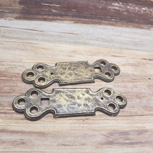 May include: Two antique brass-colored metal door hinges with decorative details. The hinges have a hammered finish and are in the shape of a rectangle with rounded edges.