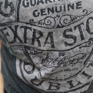K&ouml;nnte beinhalten: Ein schwarzes T-Shirt mit einem grauen und schwarzen grafischen Aufdruck eines Etiketts mit der Aufschrift "Genuine Extra Stout" und "Brewed in Ireland".