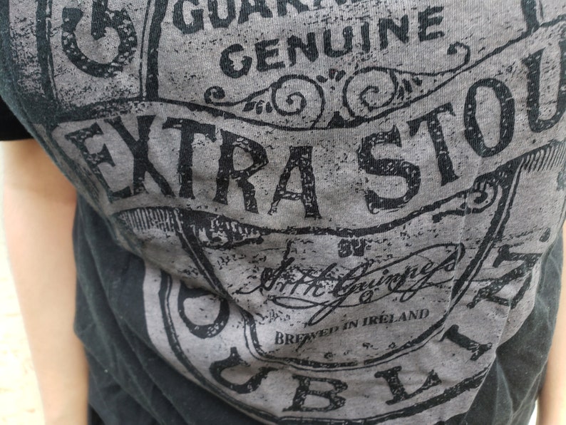 K&ouml;nnte beinhalten: Ein schwarzes T-Shirt mit einem grauen und schwarzen grafischen Aufdruck eines Etiketts mit der Aufschrift "Genuine Extra Stout" und "Brewed in Ireland".