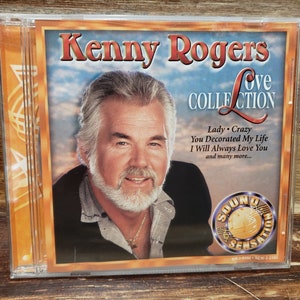 May include: A Kenny Rogers "Love Collection" CD in a clear plastic case. The album cover features a portrait of Kenny Rogers with the album title and song titles like "Lady" and "Crazy". The CD case is on a wooden surface.