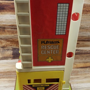 May include: A vintage Playskool Rescue Center toy building with a yellow base, red and white striped garage door, and a white ladder. The building has a sign that reads "Playskool Rescue Center" with a red cross. The garage door has a sign that reads "Engine No. 1 Do Not Block Driveway".