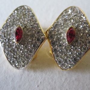 Op de afbeelding: Een paar gouden clip-on oorbellen met een rode strass in het midden van elke oorbel. De oorbellen zijn bedekt met heldere strass.